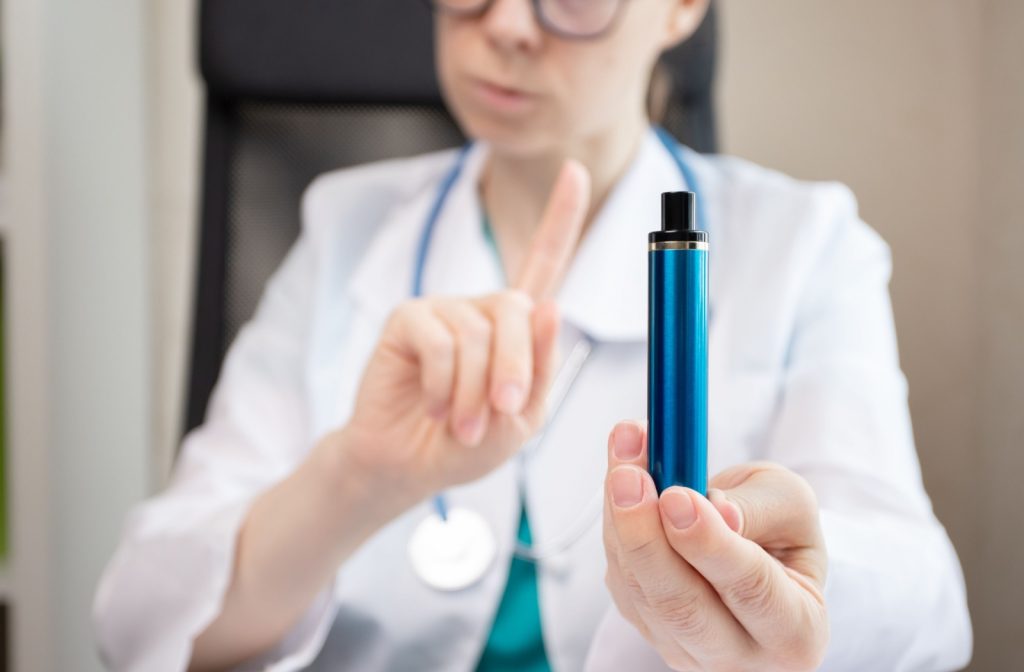 Medical professional holding a blue vape pen in one hand and raising the other hand with a warning gesture, emphasizing the health risks of vaping.
